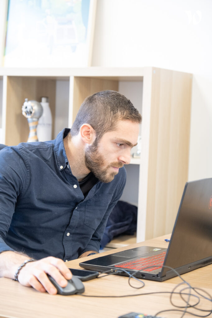 Homme concentré travaillant sur un ordinateur portable à son bureau, utilisant une souris, dans un environnement de bureau lumineux.